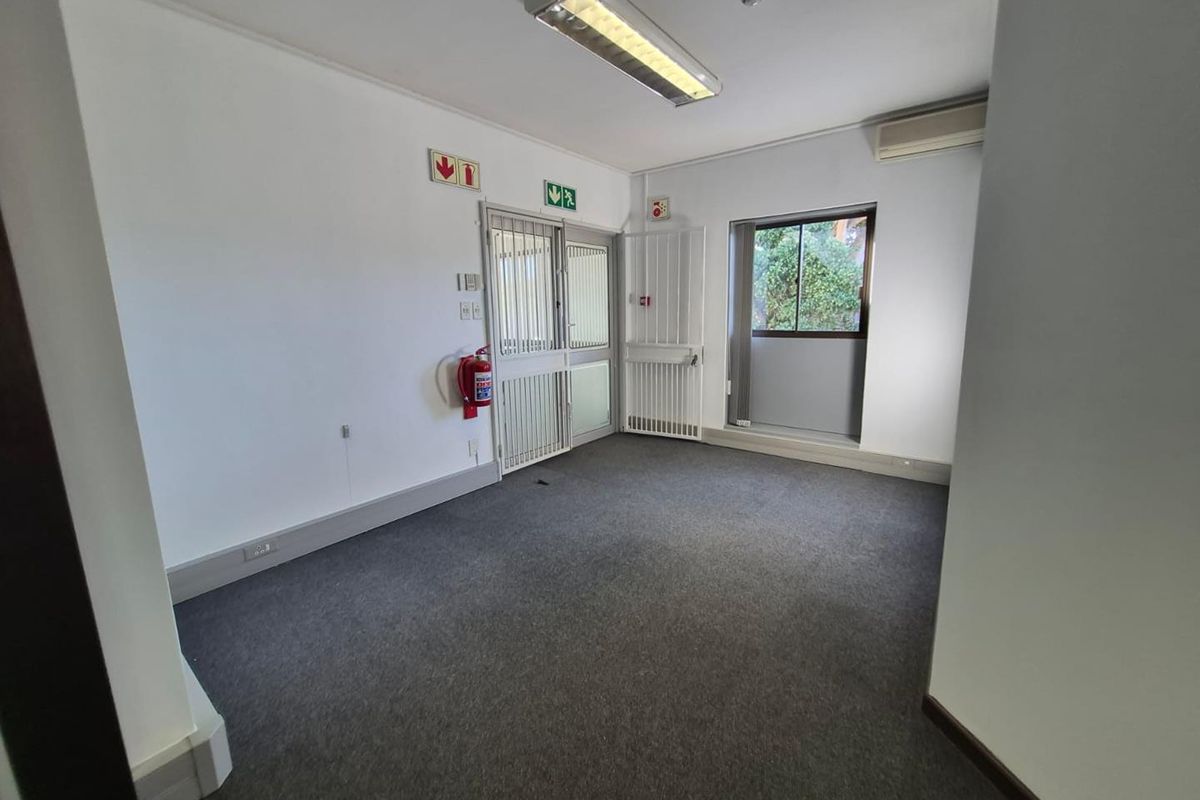 A clean, empty office space featuring carpeted flooring, a split-system air conditioner, security gates, and integrated fluorescent lighting.