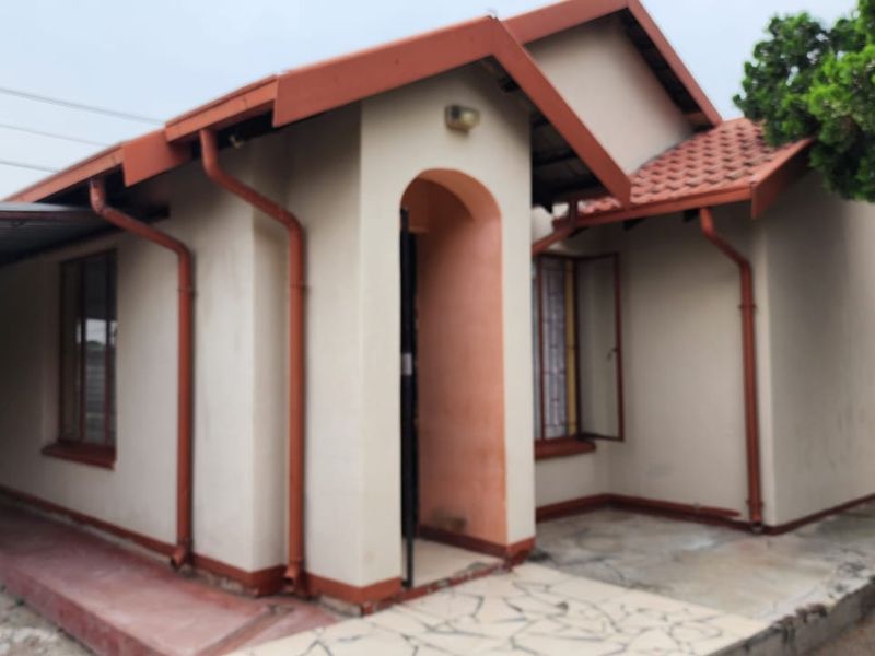Front elevation of a single-story house featuring a terracotta tiled roof, arched entrance, and attached carport with paved walkway.