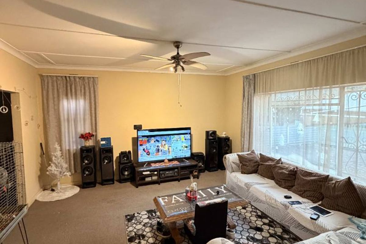 A spacious living room featuring a ceiling fan, large windows with curtains, and carpeted flooring.