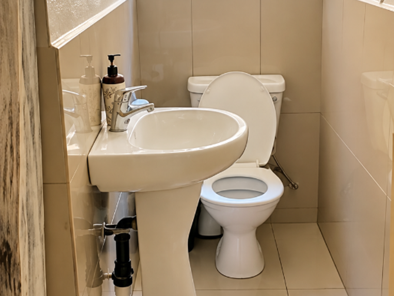 A compact bathroom featuring a pedestal basin, toilet, and neutral wall and floor tiling with a window providing natural light.