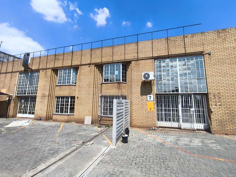 Front elevation of an industrial property featuring brick facade, steel-framed windows with security bars, and a paved driveway area.
