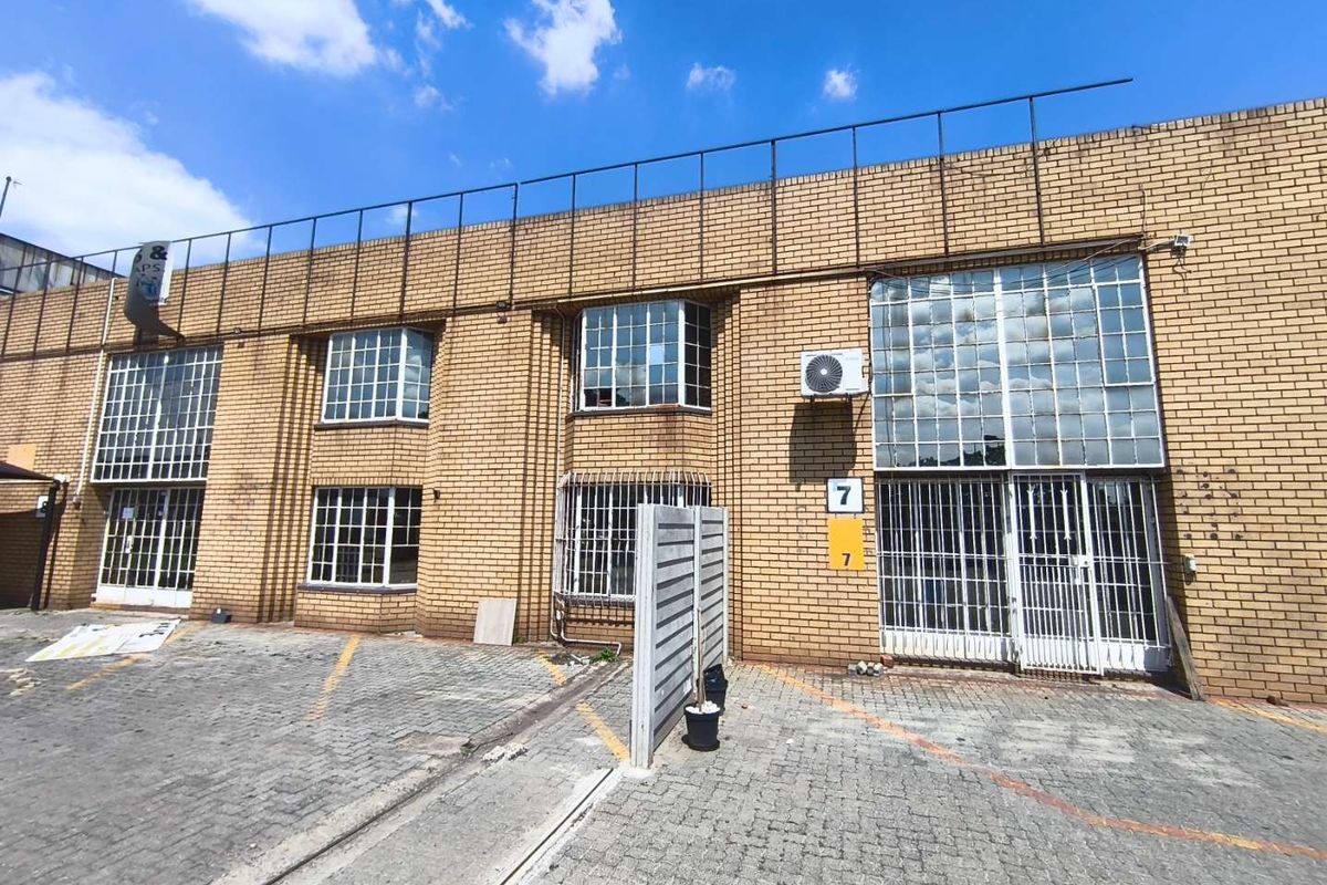 Front elevation of an industrial property featuring brick facade, steel-framed windows with security bars, and a paved driveway area.