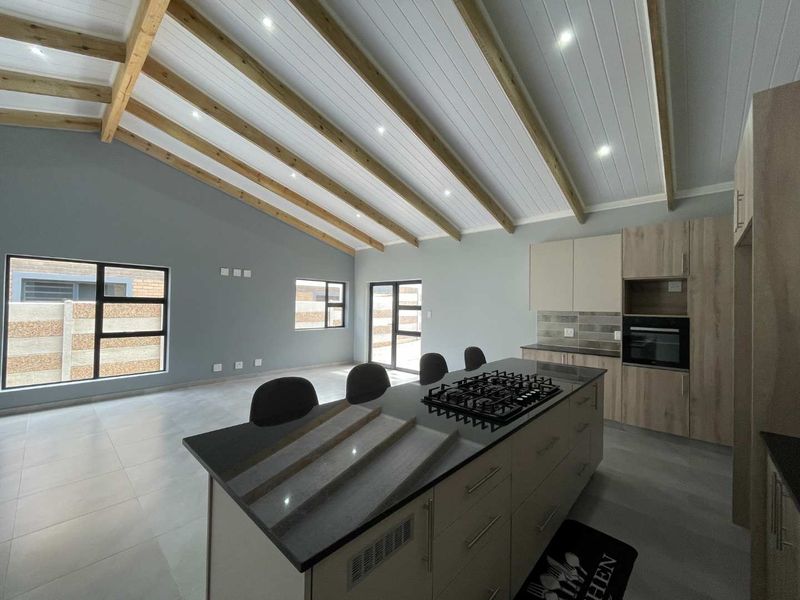 Spacious open-plan living and kitchen area featuring a vaulted ceiling with exposed timber beams, modern cabinetry, a central island with gas hob, and tiled flooring.