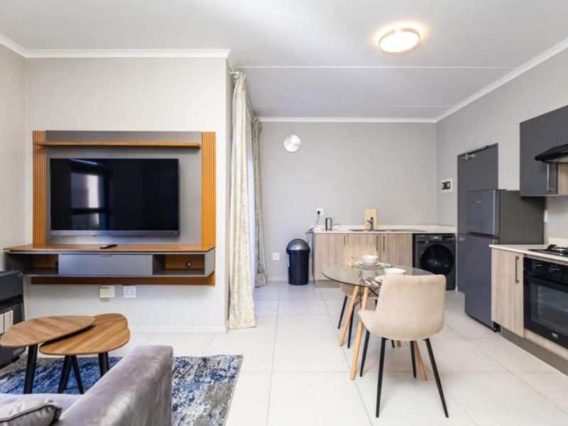 Modern open-plan living area featuring a sleek kitchen with built-in appliances and a dedicated media wall, finished with neutral tones and tiled flooring.