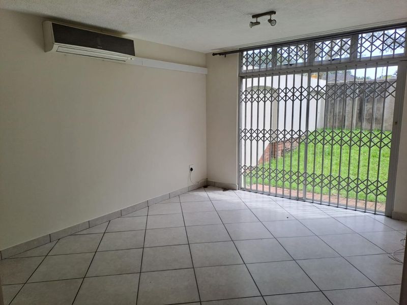 An empty living room featuring tiled flooring, a wall-mounted air conditioning unit, and a secure sliding glass door leading to the garden.
