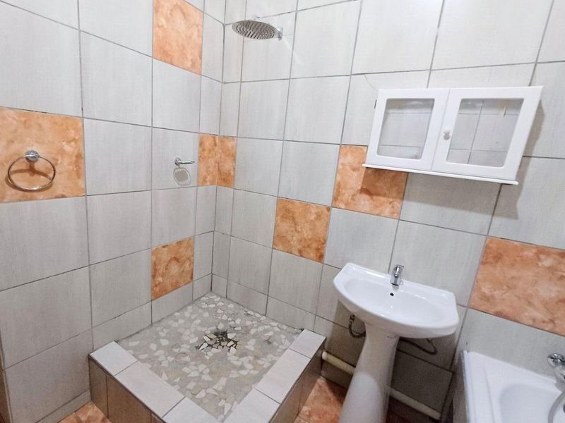 A functional bathroom featuring a pedestal basin, shower area with tiled base, and wall-mounted storage cabinet, requiring some cosmetic updates to the tiling.