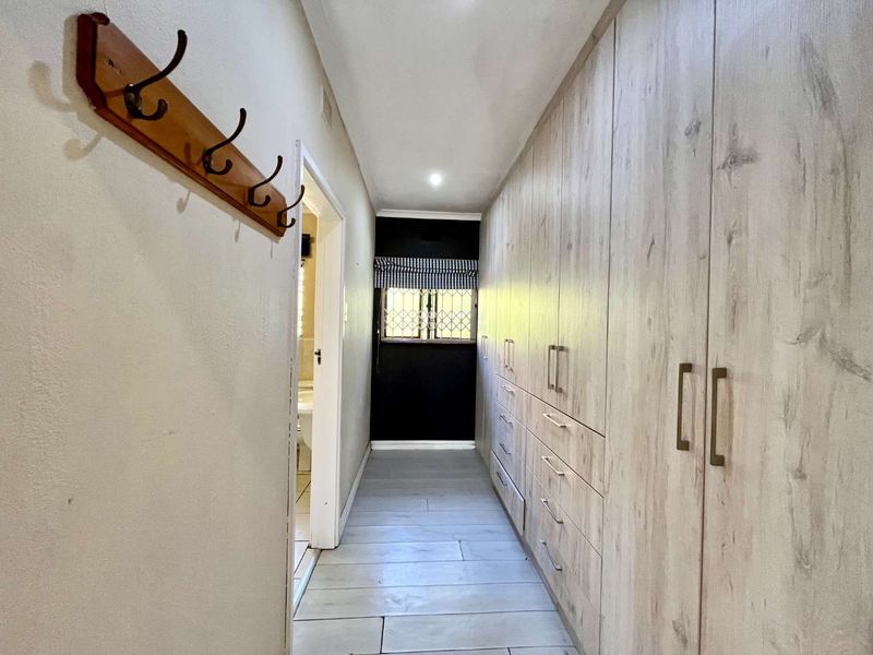 A clean and functional hallway featuring extensive built-in cabinetry with light wood-grain finish, tiled flooring, and a secure window at the end.