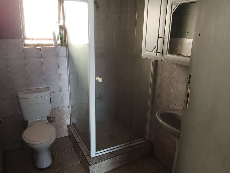 A compact bathroom featuring a shower enclosure, pedestal basin, and toilet, finished with tiled walls and flooring. Requires cosmetic updates.