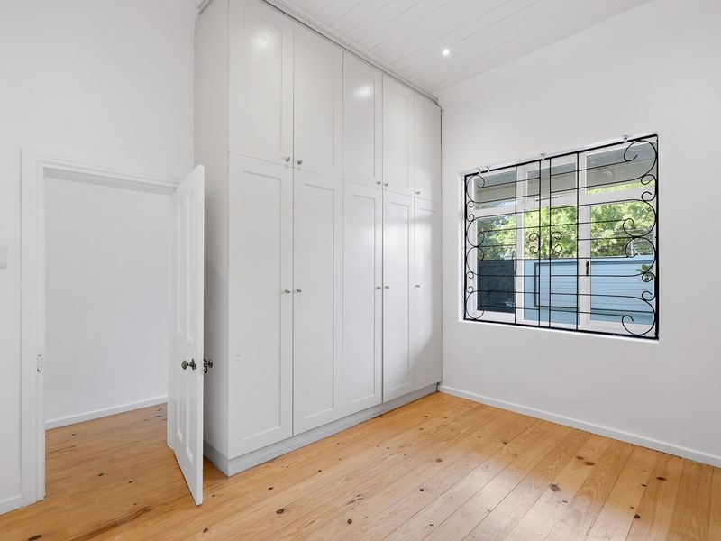 A bright, empty bedroom featuring polished timber flooring, extensive floor-to-ceiling built-in cupboards, and a window fitted with decorative security bars.