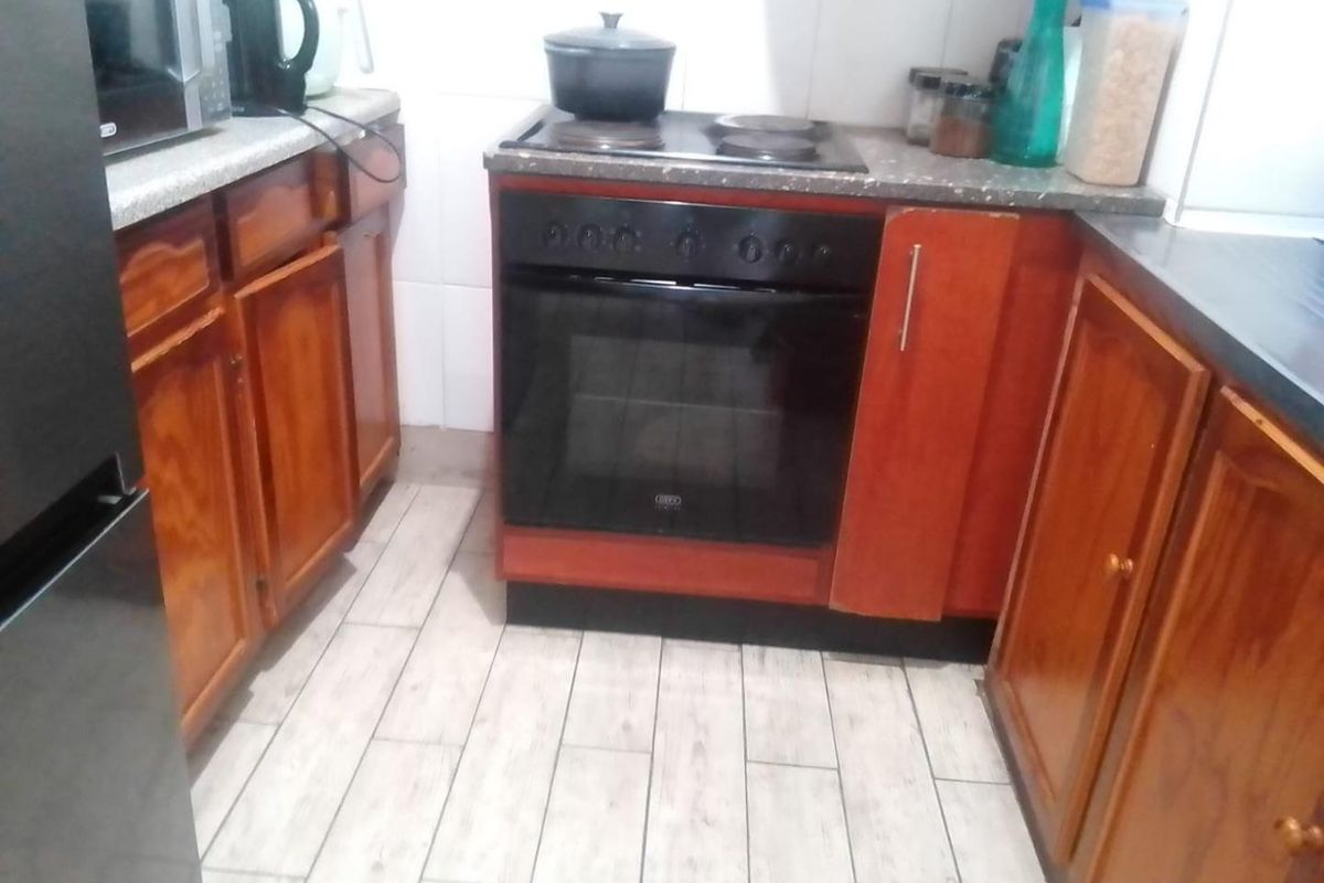 A compact kitchen featuring wooden cabinetry, stone-look countertops, a built-in oven with electric hob, and light-toned tiled flooring.