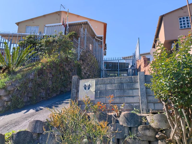 Front exterior view of the property featuring a steep driveway, security fencing, and a retaining wall structure.