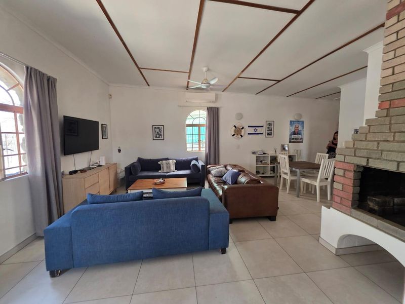 Spacious open-plan living and dining area featuring a prominent brick fireplace, tiled flooring, and an exposed beam ceiling with a ceiling fan.