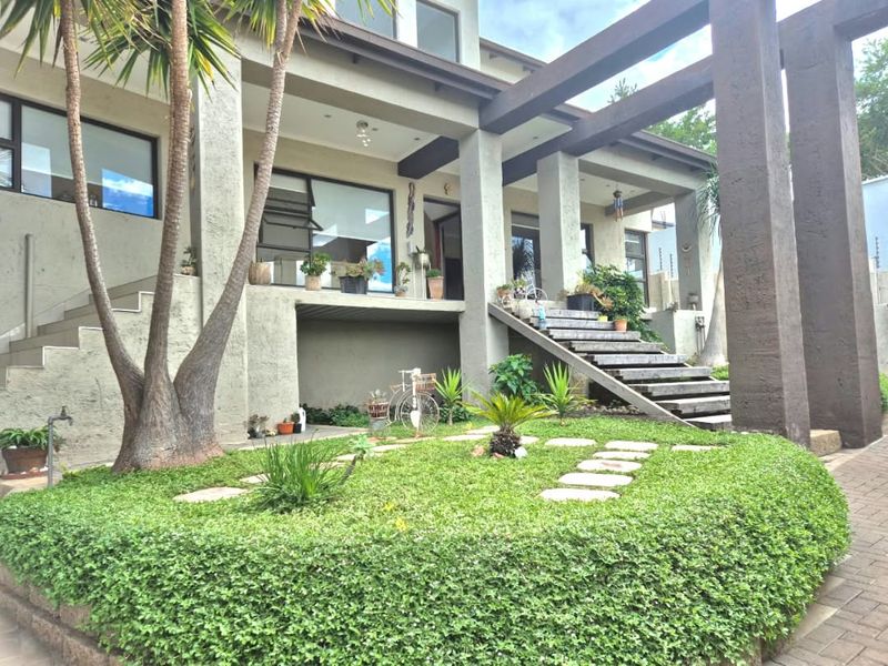 A well-maintained rear exterior featuring a raised patio, timber staircase, and a pergola structure overlooking a landscaped garden and paved driveway.