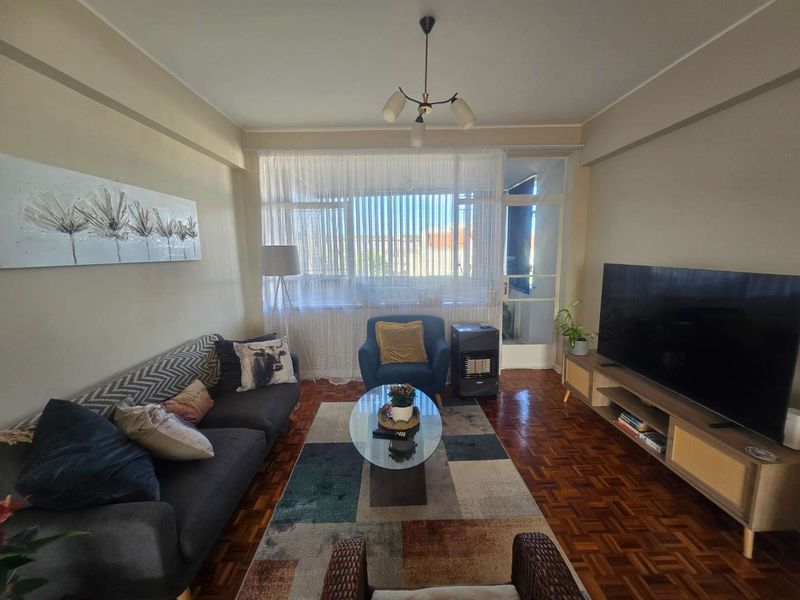 A comfortable living room featuring classic parquet flooring, natural light from a large window, and direct access to an adjoining balcony.