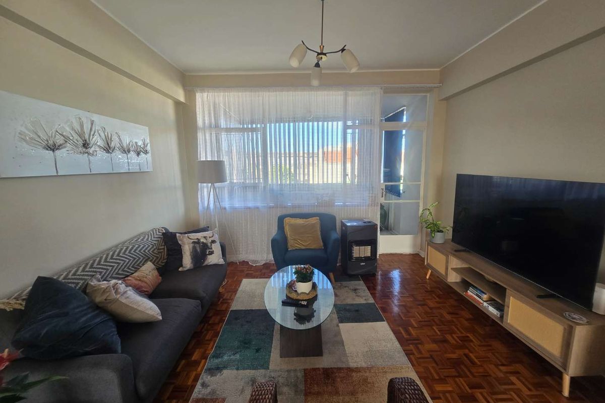 A comfortable living room featuring classic parquet flooring, natural light from a large window, and direct access to an adjoining balcony.