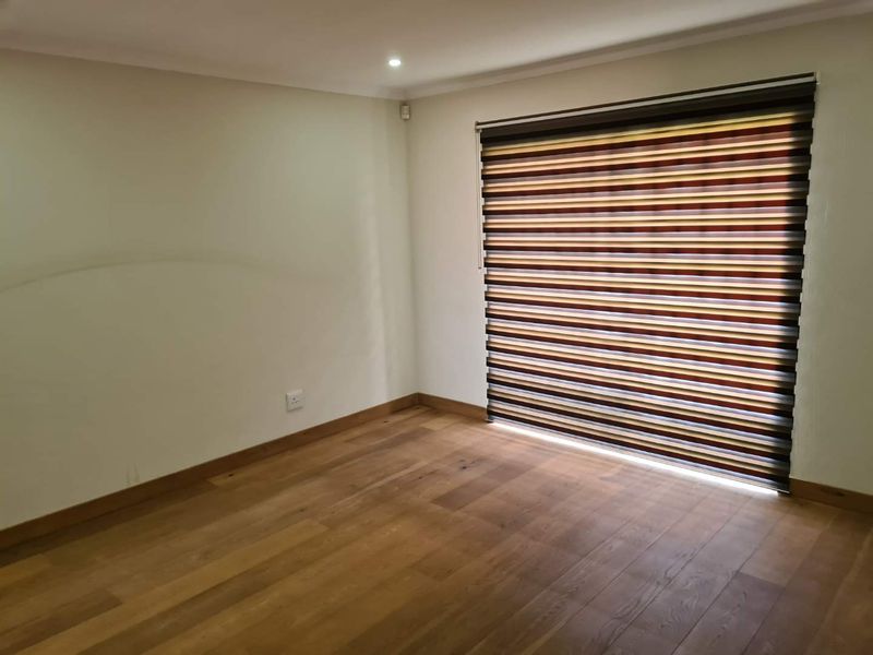 A bright, empty bedroom featuring polished hardwood flooring, neutral walls, and modern striped zebra blinds.