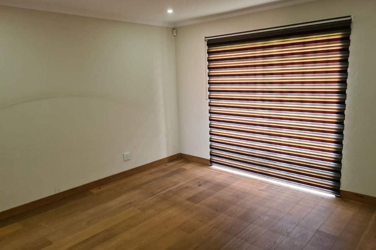 A bright, empty bedroom featuring polished hardwood flooring, neutral walls, and modern striped zebra blinds.