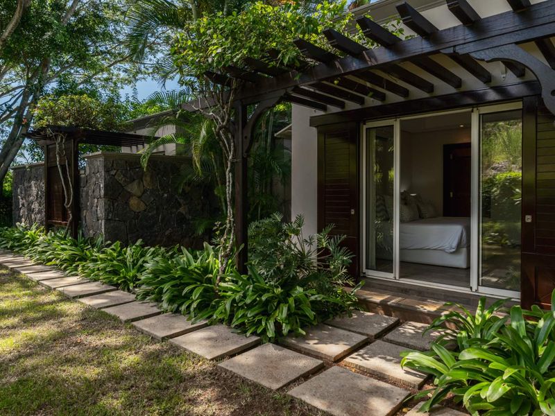 A well-maintained outdoor patio area featuring a timber pergola, stone feature wall, and paved walkway leading to a bedroom with sliding glass doors.