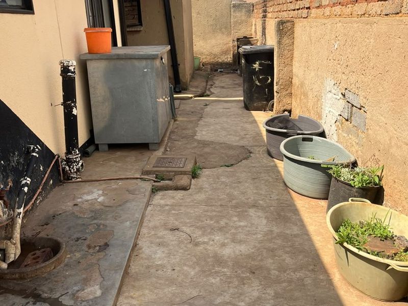 A narrow rear exterior walkway featuring a concrete path, brick boundary wall, and drainage infrastructure, showing signs of wear and weathering.