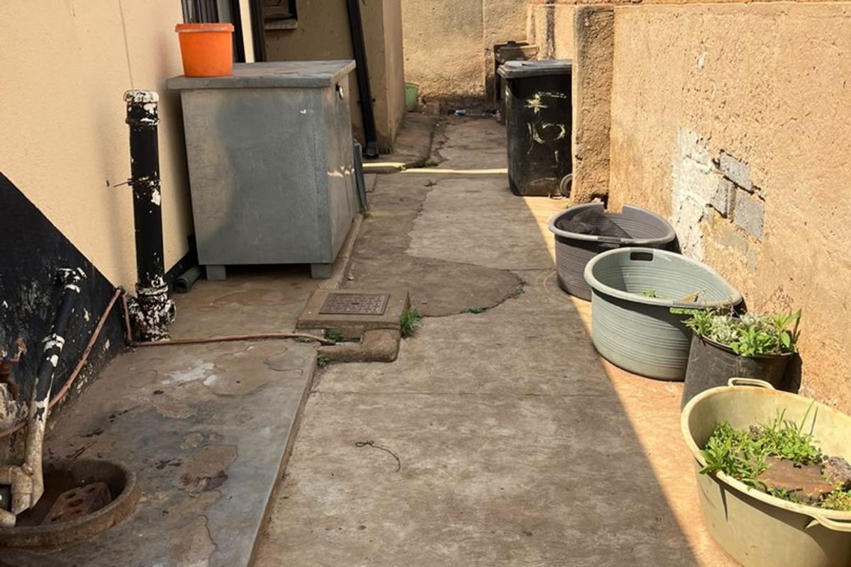 A narrow rear exterior walkway featuring a concrete path, brick boundary wall, and drainage infrastructure, showing signs of wear and weathering.