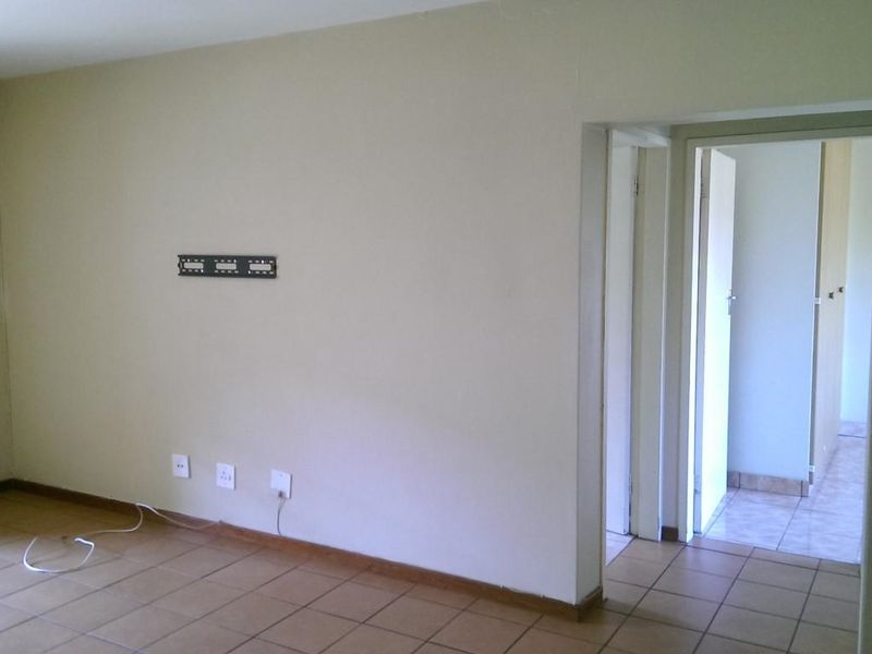 An empty living room featuring tiled flooring, wall-mounted TV bracket, and access to an adjacent hallway.