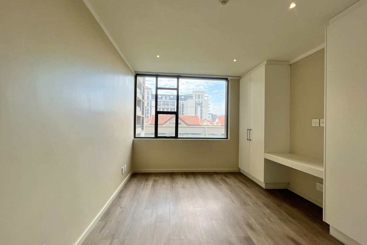 A bright, empty bedroom featuring modern wood-look flooring, built-in cabinetry with an integrated desk, and large windows providing natural light.