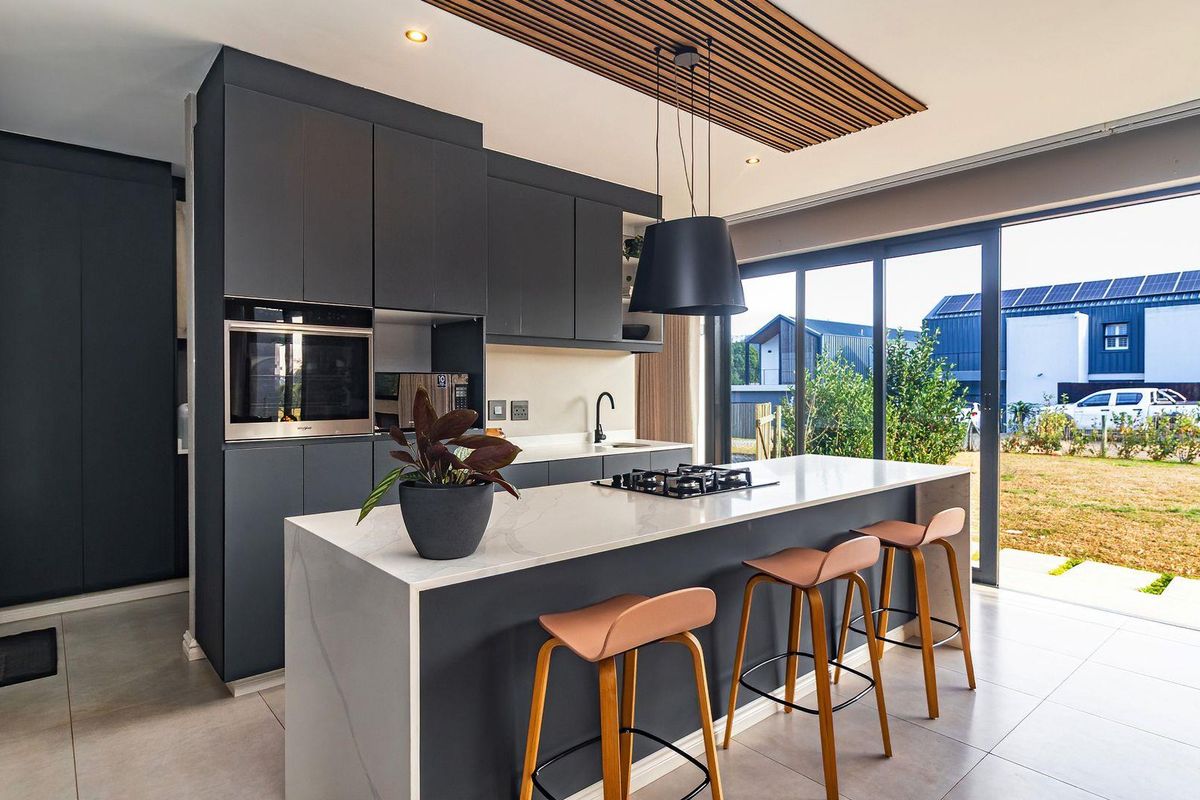 Modern open-plan kitchen featuring dark cabinetry, stone countertops, a central island with a built-in gas hob, and large sliding doors opening to the garden.