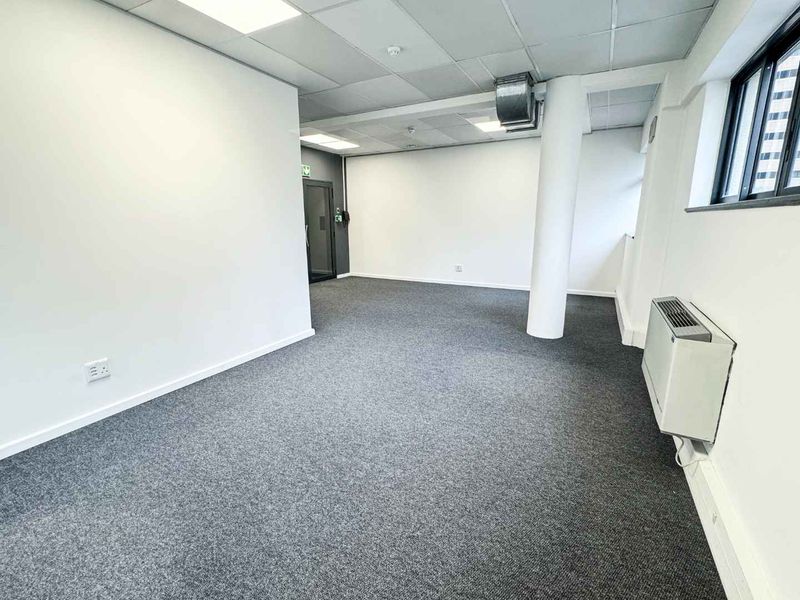 A clean, open-plan office space featuring neutral carpet flooring, a suspended ceiling with recessed lighting, and large windows providing natural light.