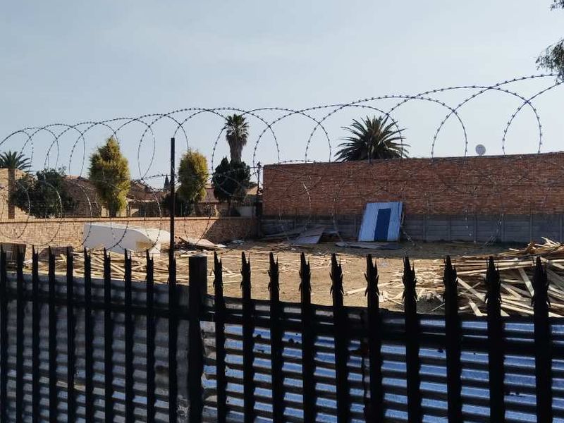 A vacant residential plot enclosed by a brick perimeter wall and metal security fencing topped with razor wire.