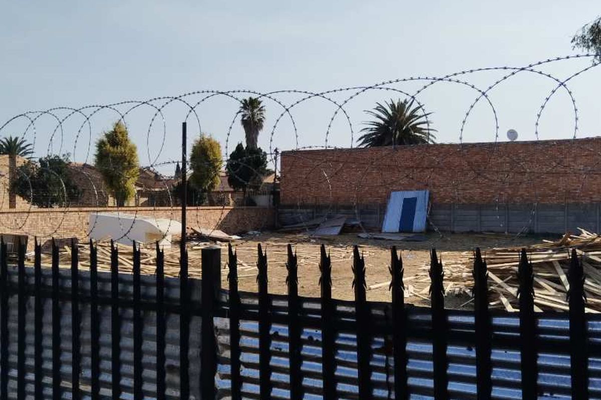 A vacant residential plot enclosed by a brick perimeter wall and metal security fencing topped with razor wire.