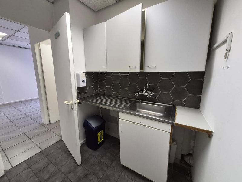 A compact office kitchenette featuring a stainless steel sink, hexagonal tile backsplash, and white cabinetry with tiled flooring.