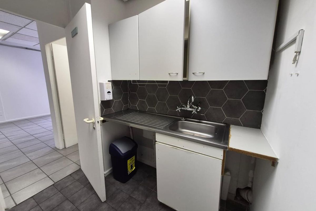 A compact office kitchenette featuring a stainless steel sink, hexagonal tile backsplash, and white cabinetry with tiled flooring.