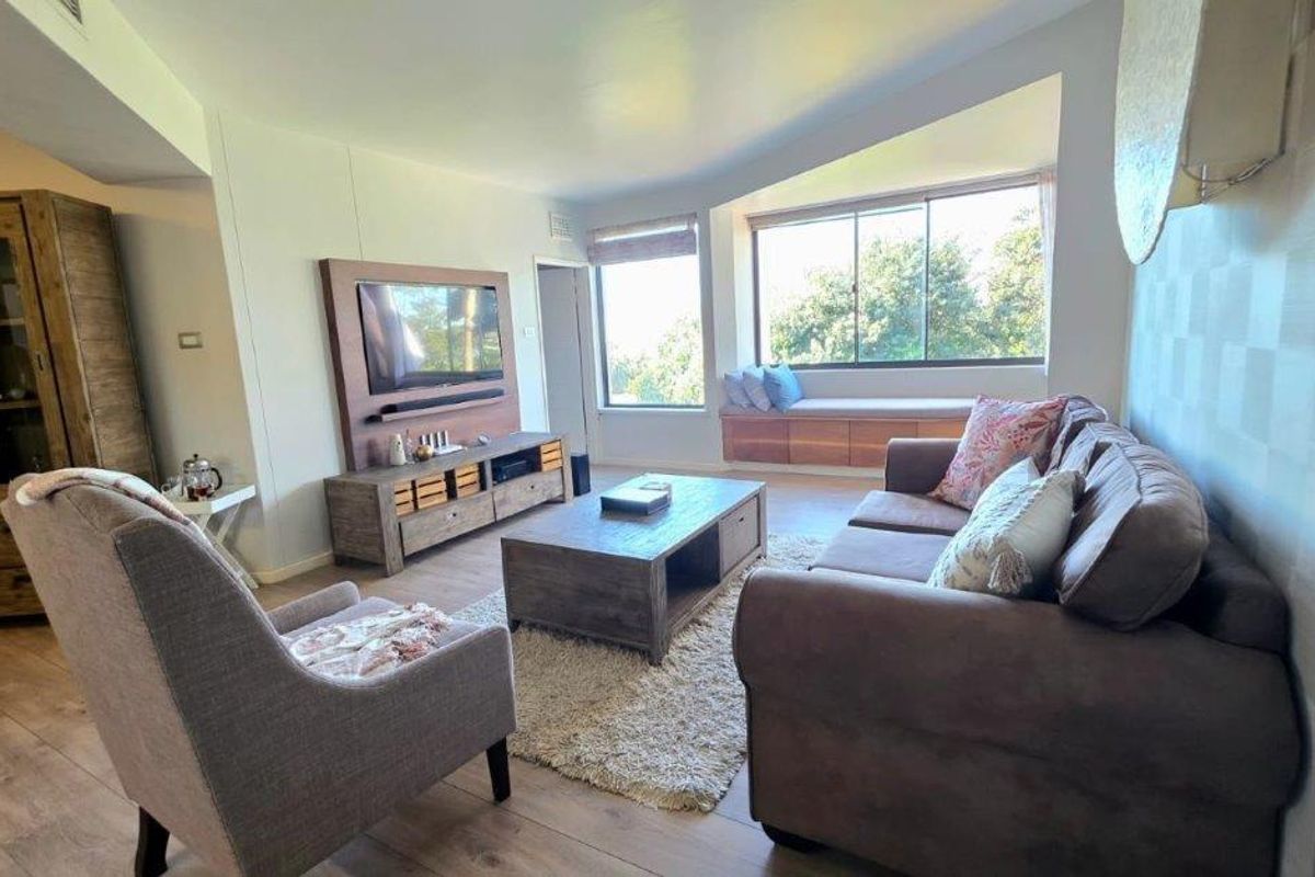 A comfortable living room featuring laminate flooring, a built-in window seat with storage, and large windows providing natural light and garden views.