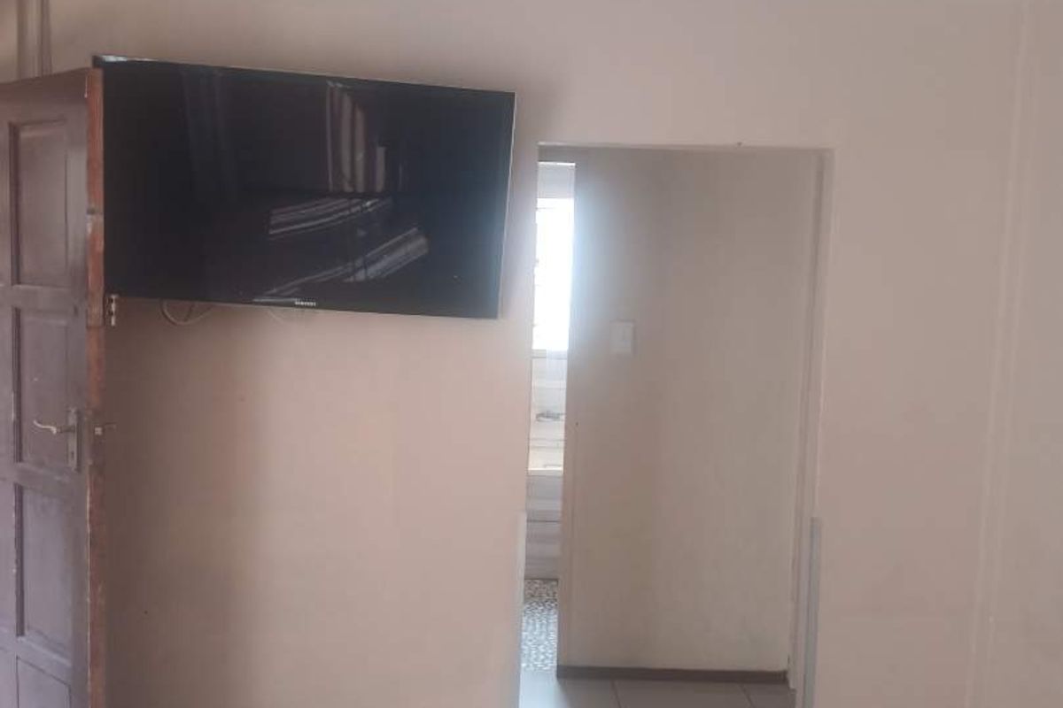 A simple living area featuring tiled flooring and a wall-mounted television, with an open doorway leading to an adjacent room.
