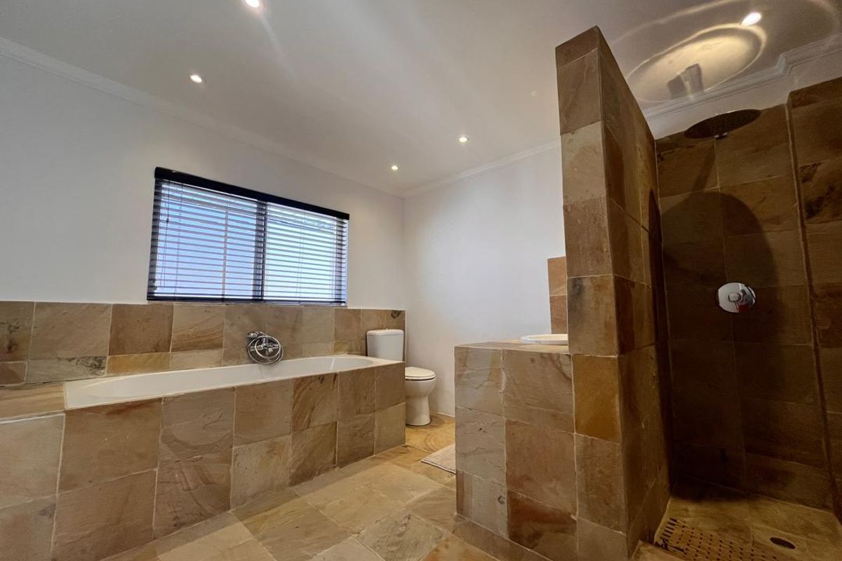 A spacious bathroom featuring natural stone tiling, a built-in bathtub, a walk-in shower with a rainfall showerhead, and recessed ceiling lighting.