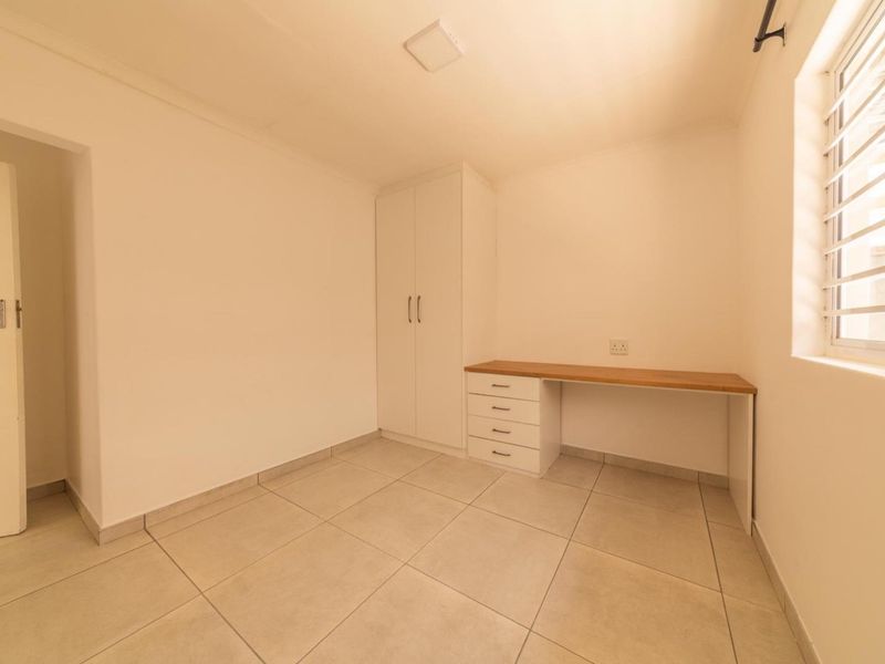 A clean, empty bedroom featuring neutral tiled flooring, a built-in wardrobe, and a functional built-in desk area with drawers.