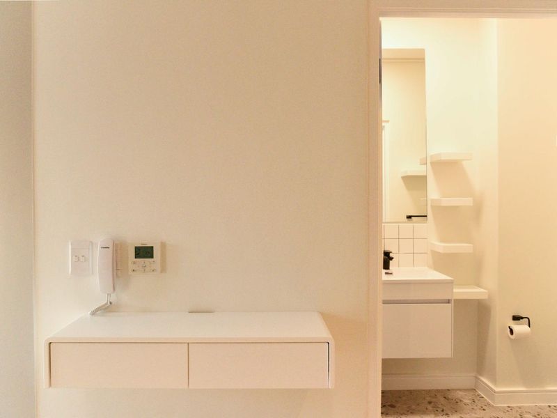 A clean, modern hallway featuring a wall-mounted console, intercom system, and a view into a bright, minimalist bathroom with built-in vanity and shelving.