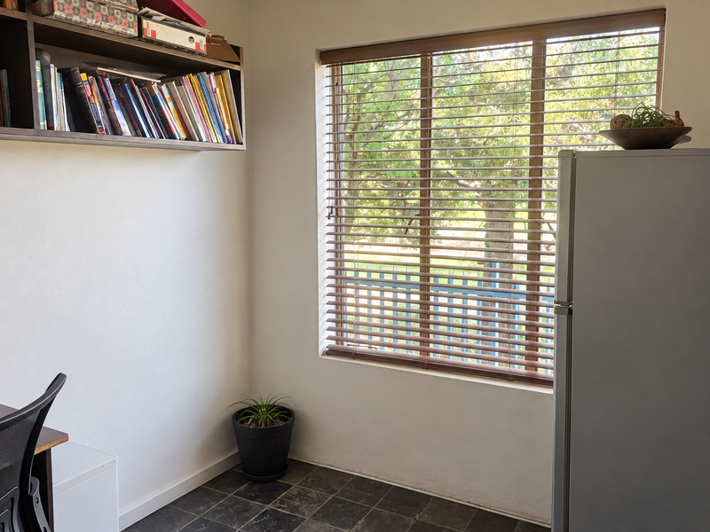 A functional home office space featuring built-in wall shelving, tiled flooring, and a large window with fitted venetian blinds.