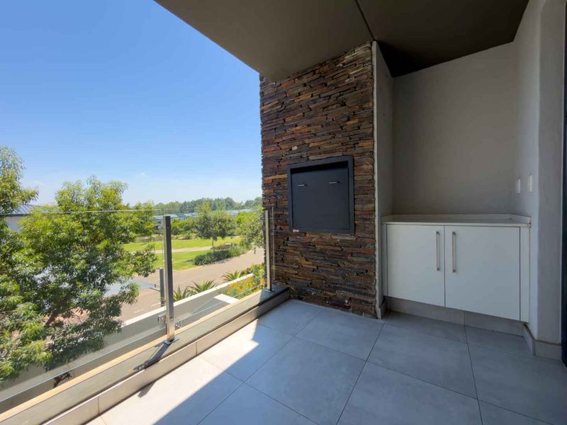 A modern covered balcony featuring a built-in braai, stone cladding, sleek white cabinetry, and a glass balustrade overlooking the surrounding landscape.