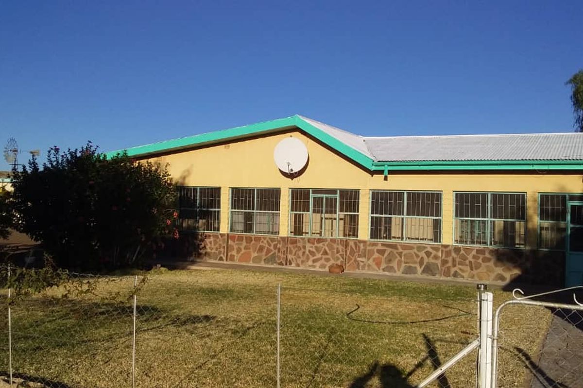A single-story farm house exterior featuring a corrugated metal roof, stone base cladding, and large windows with security bars, set within a garden.