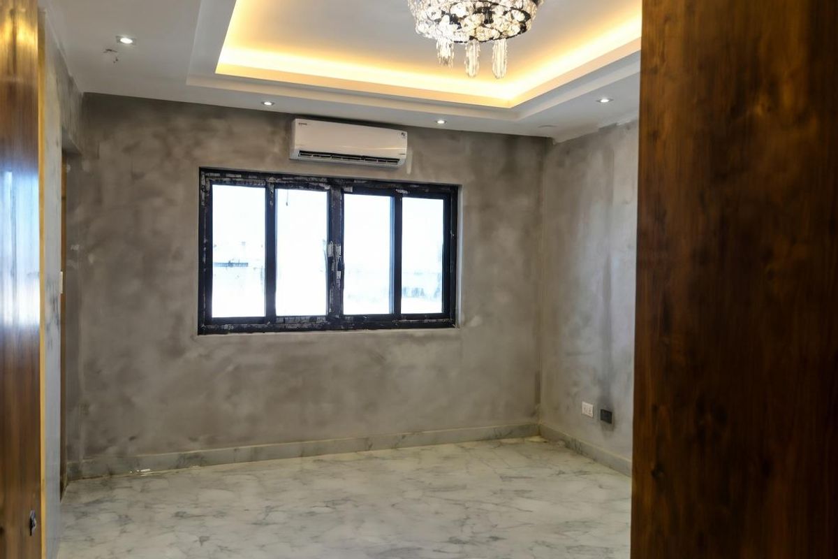 A modern, empty bedroom featuring polished marble flooring, a recessed tray ceiling with ambient lighting, a crystal chandelier, and a wall-mounted air conditioning unit.
