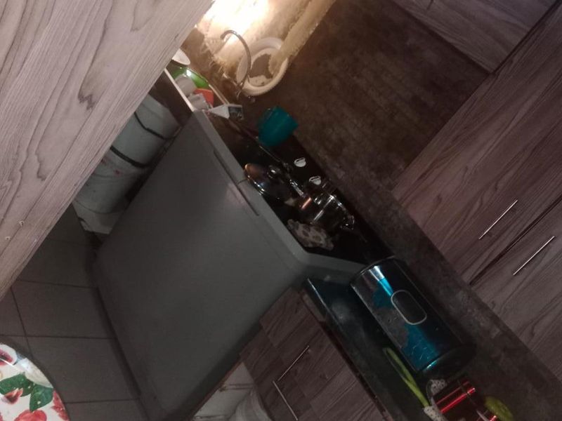 A compact kitchen featuring wood-grain cabinetry, tiled backsplash, and a stainless steel sink, currently showing signs of wear and missing cabinet components.