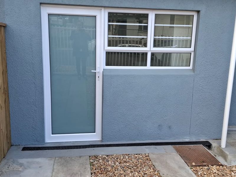 Rear exterior view of the apartment featuring a frosted glass door, aluminium window with security bars, and a paved walkway with drainage.