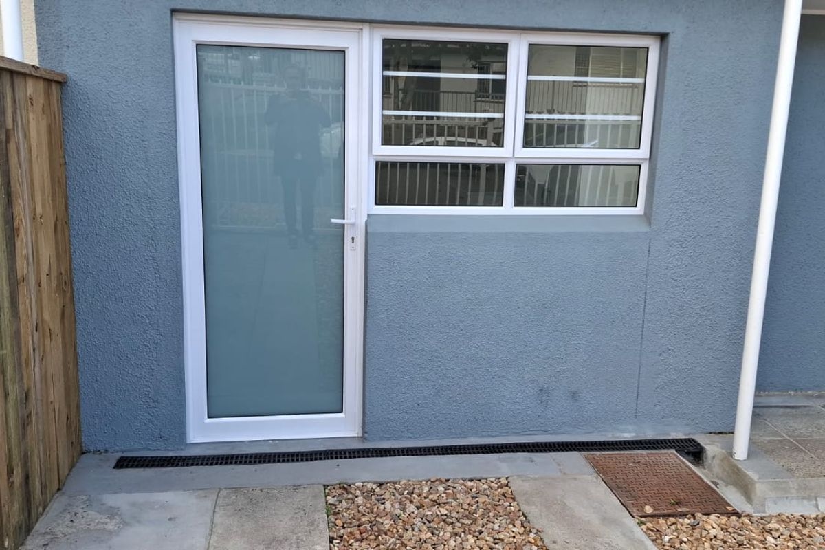 Rear exterior view of the apartment featuring a frosted glass door, aluminium window with security bars, and a paved walkway with drainage.