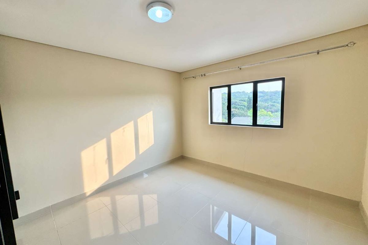 A bright, empty bedroom featuring neutral tiled flooring, a large window with exterior views, and a simple ceiling-mounted light fixture.