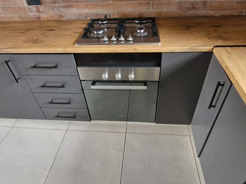 Modern kitchen featuring grey cabinetry, timber laminate countertops, an exposed brick splashback, and a built-in gas hob with oven.