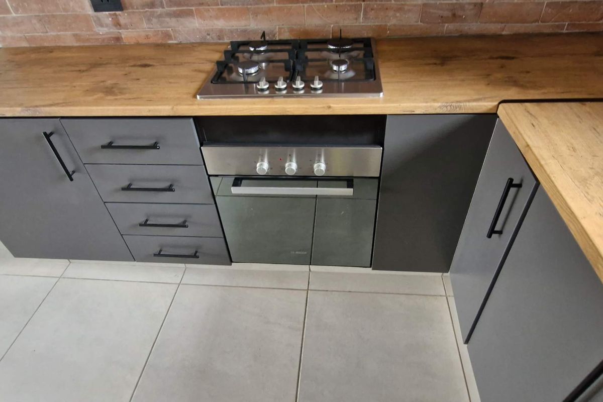 Modern kitchen featuring grey cabinetry, timber laminate countertops, an exposed brick splashback, and a built-in gas hob with oven.