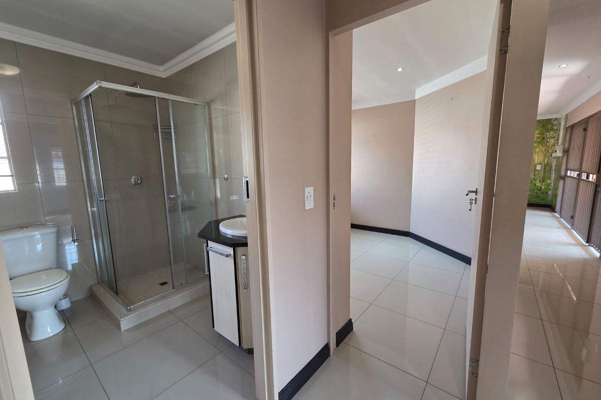 A clean, modern bathroom featuring a glass shower enclosure, vanity unit, and neutral ceramic tiling, adjacent to an open-plan office space.