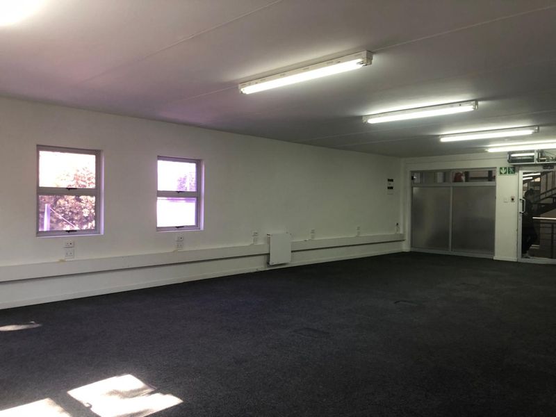 A spacious, empty office area featuring neutral walls, carpeted flooring, fluorescent lighting, and large windows providing natural light.