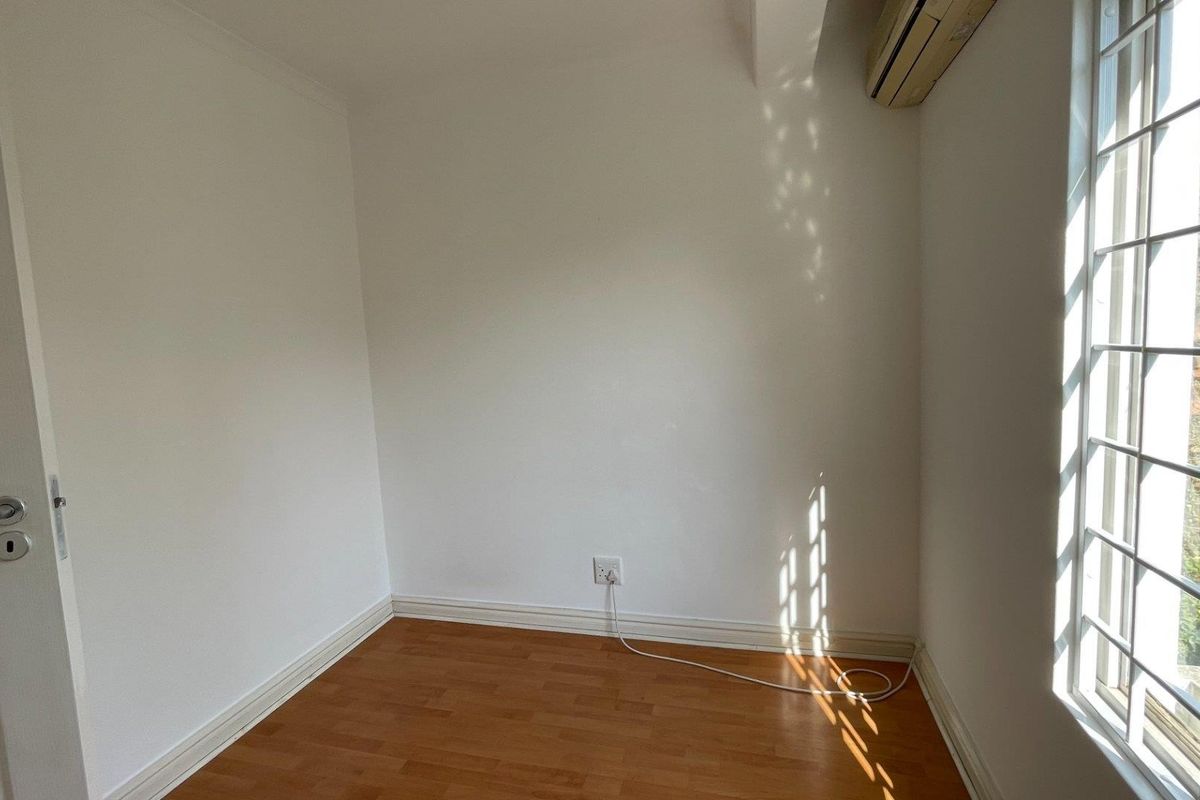 A bright, empty bedroom featuring laminate flooring, a wall-mounted air conditioning unit, and a window fitted with security bars.